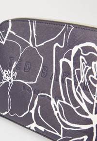Lilla stof clutch med hvidt blomstermotiv, præget "TED BAKER LONDON" logo og guldfarvet lynlåsdetaljer. Struktureret overfladefinish.
