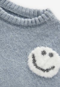 Pulover tricotat de culoare albastru deschis cu guler ribstit. Prezintă un grafic texturat cu fața zâmbitoare albă și accente gri. Aspect moale și cald.