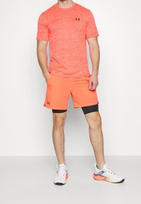 Herrträningsoutfit bestående av en ljusorange texturerad tröja och matchande orange shorts med svarta detaljer, ihop med flerfärgade löparskor.