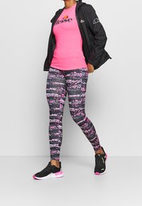 Camiseta ajustada rosa con logo, chaqueta ligera negra, leggings estampados en rosa y negro, y zapatos deportivos negros con detalles en rosa.