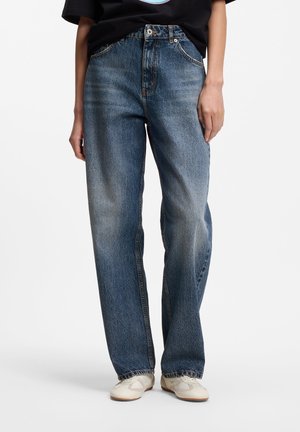 Personne portant un jean bleu délavé ample associé à des baskets blanches, debout devant un fond blanc uni.