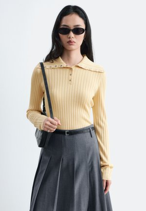 Femme portant des lunettes de soleil noires, un haut jaune côtelé à manches longues avec des boutons, une jupe plissée grise et un sac à bandoulière noir.