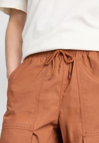 Pantalones marrones con cintura elástica y cordón de ajuste, con bolsillos laterales, textura suave y detalles cosidos a lo largo de las costuras.