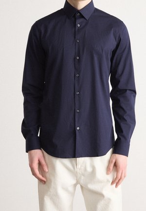 Man draagt een nauwsluitend, donkerblauw overhemd met lange mouwen en kraag, gecombineerd met crèmekleurige broek, staand tegen een effen achtergrond.