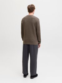 Hátrafelé néző férfi, texturált barna pulóvert, bő szürke nadrágot és fekete cipőt visel, egyszerű fehér háttér előtt.