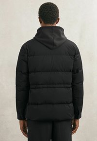 Persoon gezien van achteren, gekleed in een zwarte gewatteerde jas met capuchon en lange mouwen tegen een effen lichte achtergrond.