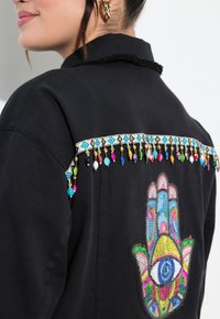 Mustapaita, jossa on monivärinen brodeeraus kädestä, jossa on silmä. Koristelu nauhat ja helmet eri väreissä hartiasaumalla.