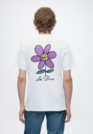 Ung person med krøllet hår iført hvid T-shirt med stor lilla blomst med basketballcenter og teksten "Les Deux" på ryggen.