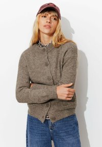 Cardigan en maille marron avec un col rond, fermeture par boutons, poignets et ourlet côtelés. Porté sur une chemise à carreaux, associé à un jean bleu.