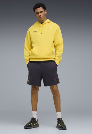 Uomo in piedi con le mani nelle tasche, indossa una felpa gialla Puma, pantaloncini scuri con stampa di Pikachu, calzini bianchi e scarpe da ginnastica nere Puma su sfondo grigio.