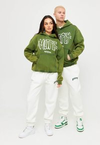 Due modelli che indossano felpe verdi coordinate con il testo "Multiply Apparel" e pantaloni della tuta bianchi, in piedi davanti a uno sfondo bianco semplice.