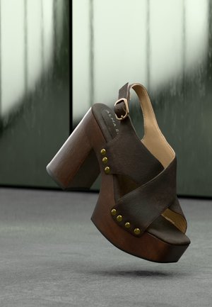 Sandale plateforme en cuir marron avec talon carré en bois, clous dorés et bride de cheville avec boucle, présentée sur un fond flou.