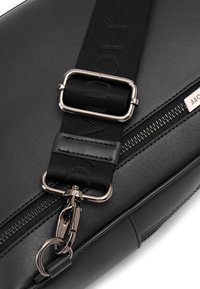 Schwarze Leder-Crossbody-Tasche mit einem breiten Trageriemen, der mit eingravierten Buchstaben verziert ist, metallischen Beschlägen und einem Reißverschlussdetail. Glatte Textur.