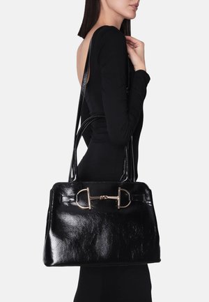 Donna con abito nero a manica lunga che porta una grande borsa in pelle lucida nera con dettagli in metallo dorato a forma di morsetto per cavallo sulla tracolla.