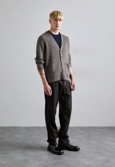 Grå ribbad cardigan med framfickor, bärs över en marinblå t-shirt, tillsammans med svarta byxor och skor. Modellen står på en grå bakgrund.