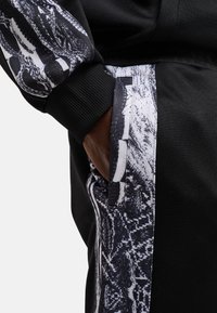 Sorte joggers med et stort, tekstureret slangeprintdetalje på siden. Stoffet er blødt med ribbede manchetter og en sidelomme.