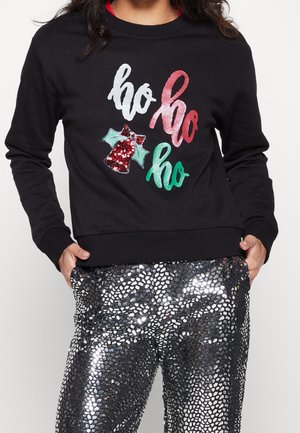 Schwarzer Pullover mit dem Schriftzug "ho ho ho" in Weiß, Rot und Grün sowie einer roten, paillettenbesetzten Glocke, kombiniert mit glänzenden silbernen Hosen im Schlangenledermuster.