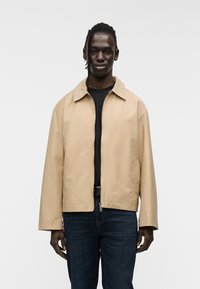 Μπεζ ζακέτα με φερμουάρ και απλωτό γιακά, με χαλαρή εφαρμογή και ορατές ραφές. Φορεμένη πάνω από μαύρο τοπ με σκουρόχρωμο μπλε τζιν.