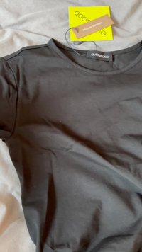 Tricou negru din bumbac, cu mâneci scurte, textură netedă, gât rotund și umeri ușor structurați. Etichetele atașate includ informații despre marcă.
