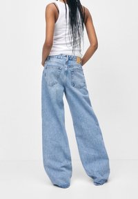 Jeans de pierna ancha de color azul claro, hechos de tela vaquera, con bolsillos traseros y un parche de cuero marrón en la cinturilla. Combinados con una camiseta sin mangas blanca.