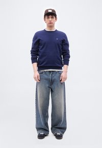 Νέος άντρας που στέκεται, φοράει μπλε ναυτικό φούτερ, καφέ καπέλο, φαρδιά ξεθωριασμένα τζιν και σκούρα αθλητικά παπούτσια, με φόντο απλό ανοιχτό χρώμα.