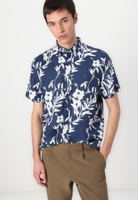 Camisa de manga corta hecha de tela azul marino con patrones florales blancos, cuello con botones y un pequeño detalle de logo en el pecho. Pantalones marrones visibles.