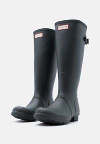 Ψηλές μαύρες λαστιχένιες μπότες βροχής με υφασμένες σόλες και κόκκινο λογότυπο Hunter κοντά στην κορυφή, μία μποτά μπροστά ελαφρώς γερμένη σε λευκό φόντο.