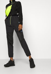 Pantalones deportivos negros con paneles de malla, cintura elástica con cordón y un corte ajustado; combinados con una chaqueta negra con cremallera y una bolsa verde neón.