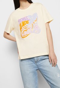 Joven mujer vestida con una camiseta color crema con un texto y diseño en espiral de "Paz y Amor" en púrpura y naranja, combinada con unos jeans rotos de color azul claro.