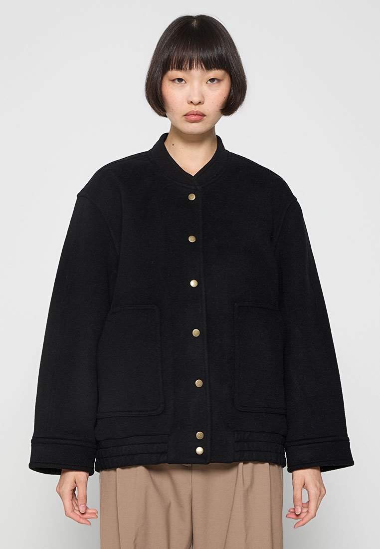 Schwarze Fleecejacke mit rundem Kragen, goldenen Druckknöpfen und zwei vorderen Taschen. Das Design ist oversized mit einer geraden Form.