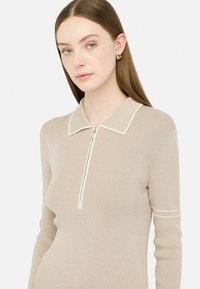Maglione a costine beige con colletto zip, dotato di rifiniture bianche e maniche lunghe. Texture liscia, design aderente e dettagli minimi.