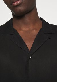 Chemise noire à boutons avec un col légèrement brillant. Le tissu est lisse avec une texture subtile, présenté en gros plan.