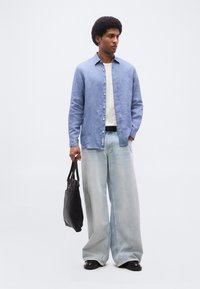 Άνδρας που φοράει ανοιχτό μπλε πουκάμισο με κουμπιά, λευκό εσώρουχο, ανοιχτόχρωμα τζιν με φαρδιά πόδια, μαύρα παπούτσια και κρατάει μαύρη τσάντα tote, όρθιος σε λευκό φόντο.