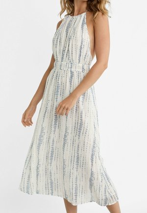 Vrouw draagt een mouwloze witte midijurk met verticale blauwe stippellijnen en een halternek, staand tegen een effen achtergrond.