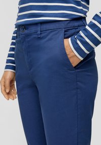 Dunkelblaue Hosen aus glattem Material, mit einer klassischen Knopfschließe und seitlichen Taschen. Kombiniert mit einem gestreiften Langarmhemd.