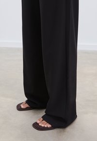 Zwarte, wijde broek van soepel materiaal, gecombineerd met donkerbruine instap sandalen, waarbij blote tenen zichtbaar zijn op een neutrale vloer.