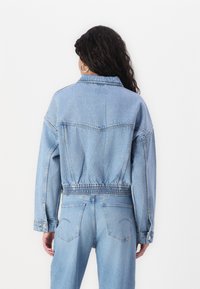 Kortad denimjacka i ljusblått, med elastisk midja, klassisk krage och synliga sömnadsdetaljer på ryggen och ärmarna.
