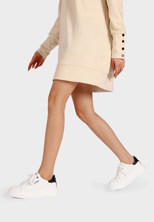 Sneakers bianchi con accento nera sul tallone, design con lacci e tessuto testurizzato. Indossati con un abito beige, evidenziando lo stile delle calzature.