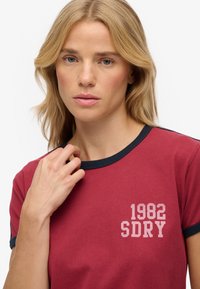 Camiseta de algodón roja con detalles en azul marino en el cuello y los bordes de las mangas. Presenta un gráfico impreso en blanco: "1982 SDRY" en la parte delantera.