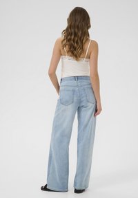 Jeans bleu clair à jambe large avec taille haute, texture décolorée et poches arrière détaillées, associés à un débardeur crème avec dentelle.