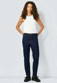 Calças jeans escuras com um corte slim e design clássico de cinco bolsos, combinadas com uma camisa branca sem mangas e sapatilhas pretas, de pé contra um fundo neutro.