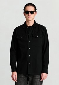 Schwarzes Langarmshirt mit zwei Brusttaschen, Knopfverschluss und glatter Textur. Kombiniert mit schwarzen Sonnenbrillen und grauen Jeans.