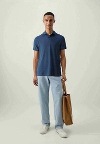 Camisa polo azul-marinho, de mangas curtas, com colarinho clássico, combinada com jeans azul claro e ténis brancos. Bolsa de mão castanha segurada na mão direita.