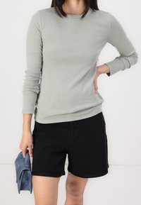 Osoba v světle zeleném žebrovaném topu s dlouhým rukávem a černých šortkách, držící malou modrou kabelku v jedné ruce a s druhou rukou na pase.