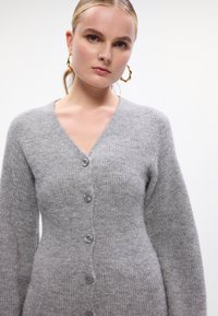 Cardigan a maglia grigia chiara con scollo a V, maniche lunghe e cinque bottoni sul davanti. Texture morbida con un motivo a coste.