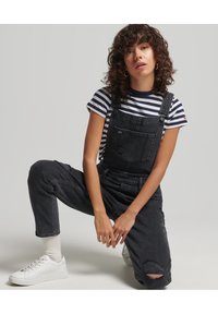 Zwarte denim overall met een verouderd detail, gedragen over een marineblauw en wit gestreept t-shirt. Gecombineerd met witte sneakers en geribde sokken.