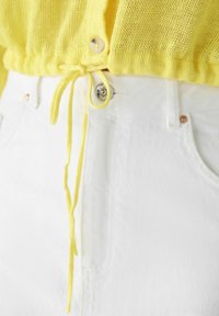 Gelbe Strickjacke mit Kordelzug in der Taille und Knopfverschluss, kombiniert mit weißen Jeans aus Denim, die über einen Metallknopf und dezente Nähte verfügen.