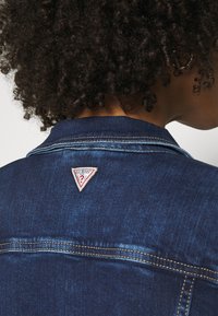 Dark blue denim jacket featuring a triangle-shaped, embroidered logo in red and white on the back. Visible stitching and texture details.