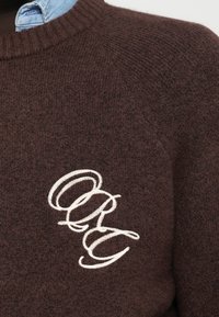 Pulover tricotat maro cu un guler rotund cu model în relief și un logo brodat alb pe față, prezentând un material texturat asemănător lânii.