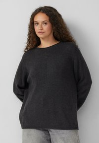 Jonge vrouw met lang krullend haar, gekleed in een losse donkergrijze trui en lichte grijze jeans, staand tegen een effen lichte achtergrond.
